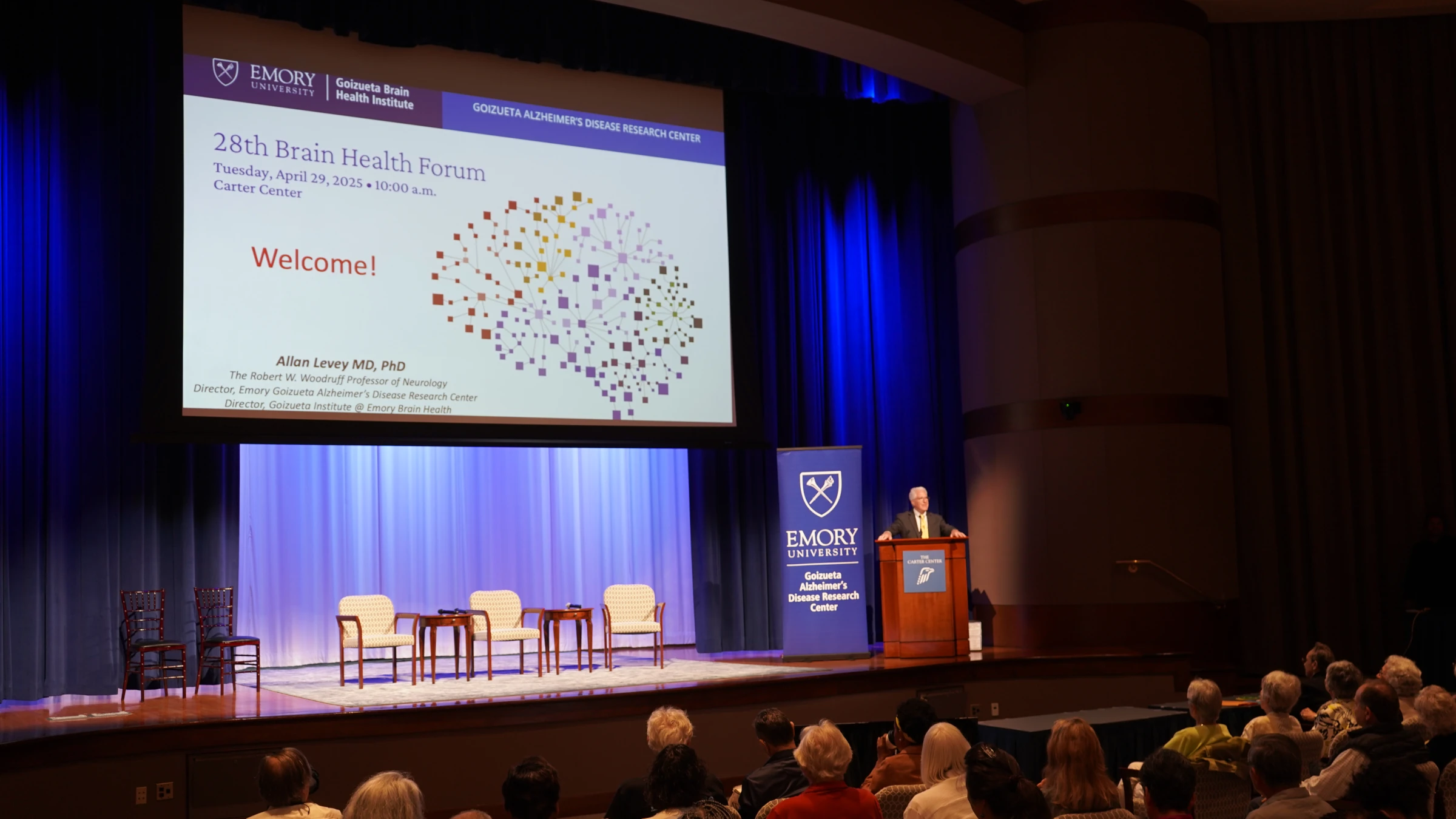 Speaker at the 28th Brain Health Forum at Emory University, standing at a podium beside a large projection screen displaying the event title, date, and welcome message in front of an audience.